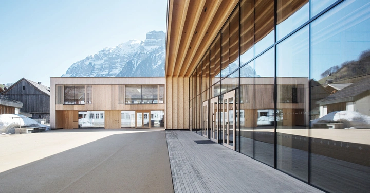 Galerie-Bild 6: Holzgebäude mit großen Glasfenstern reflektiert die Umgebung während im Hintergrund schneebedeckte Berge und traditionelle Holzhäuser zu sehen sind. Das Gebäude steht in einem weiten offenen Hof.