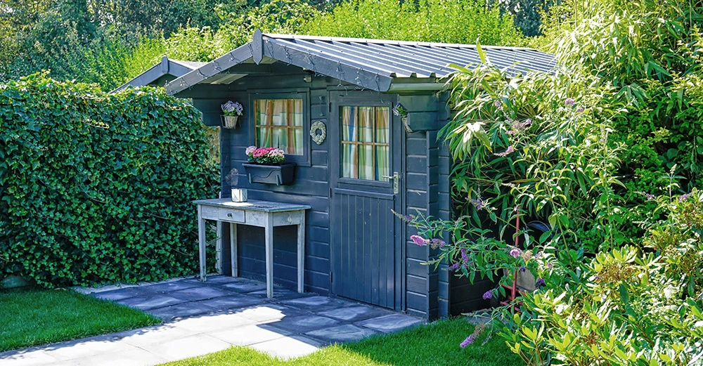 Dunkelgraue Gartenhütte mit Fenster und Topfblumen stehendes Holztischchen davor umgeben von grünem Rasen und dichten Pflanzen im Garten.