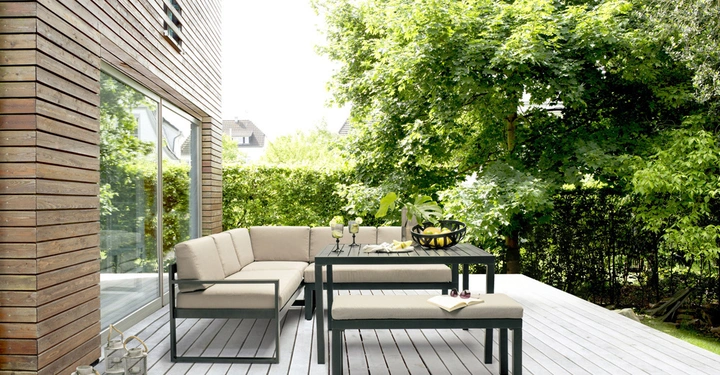 Galerie-Bild 5: Ecksofa und Esstisch mit Bänken stehen auf einer Holzterrasse neben einem modernen Haus mit Holzverkleidung umgeben von üppigem grünem Laub und Bäumen im Garten.