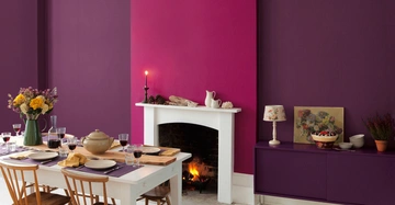 Ein gedeckter Tisch mit Blumenvasen und Geschirr vor einem violett-pink gestrichenen Raum mit einem Kamin an der Wand Dulux.