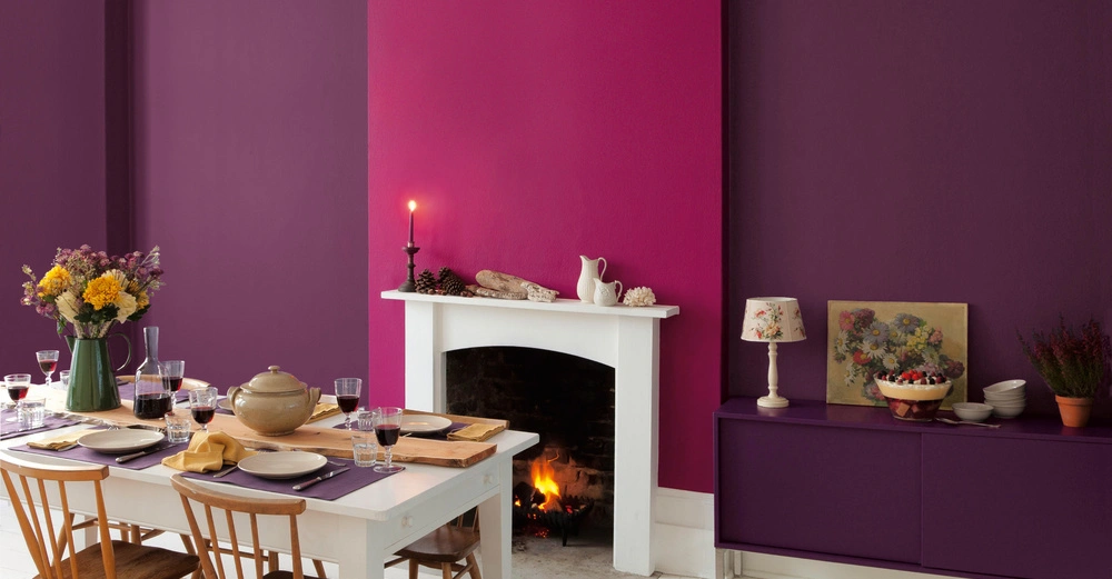 Ein gedeckter Tisch mit Blumenvasen und Geschirr vor einem violett-pink gestrichenen Raum mit einem Kamin an der Wand Dulux.
