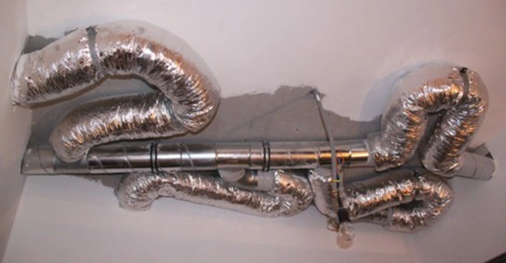 Galerie-Bild 32: Silberne Lüftungsrohre verlaufen in geschwungenen Linien unter der Decke eines Raumes, die von grauem Putz umgeben sind und teilweise freiliegende Rohrverbindungen aufweisen.