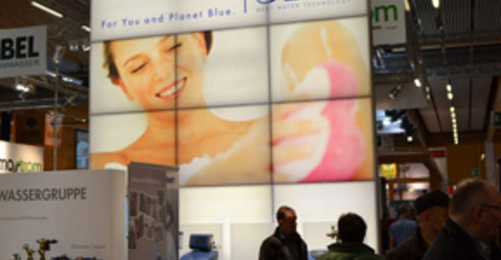 Galerie-Bild 18: Eine Frau hält einen rosa Schwamm und lächelt. Messeumgebung mit Menschen und Ständen. Schrifttext BWT For You and Planet Blue. Produkte für Wasseraufbereitung.