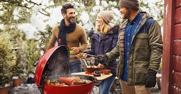 Drei Personen stehen lächelnd um einen roten Grill herum Grillen im verschneiten Außenbereich Sie tragen Winterkleidung und bereiten Essen zu