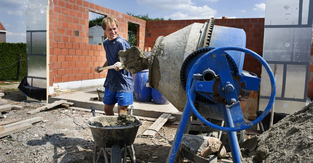 Arbeiter am Bau schaufelt Zement in blauen Betonmischer, im Hintergrund halb gemauerte Garage, orange Ziegel