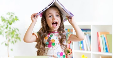 Ein Kind hält ein Buch über dem Kopf lächelnd in einem hellen Raum mit Regalen voller Bücher und Pflanzen im Hintergrund.