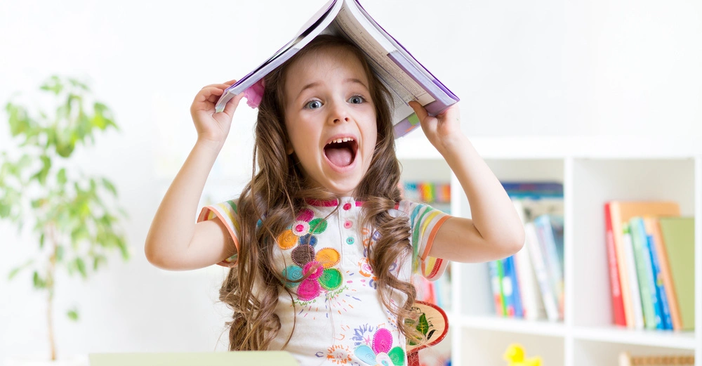 Ein Kind hält ein Buch über dem Kopf lächelnd in einem hellen Raum mit Regalen voller Bücher und Pflanzen im Hintergrund.
