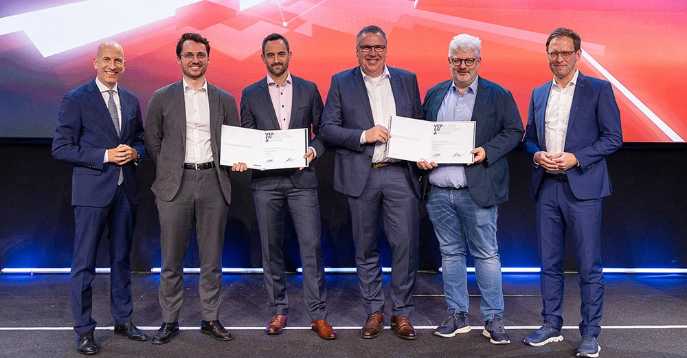 6 Männer im Anzug stehen lächelnd auf Bühne mit blau-rotem insiven Hintergrund und halten ihre Auszeichnung in die Höhe. 