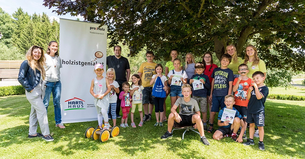 Eine Gruppe von Kindern und Erwachsenen posiert fröhlich für ein Foto auf einer grünen Wiese vor einem Poster mit der Aufschrift proHolz Niederösterreich holzistgenial.at und HARTL HAUS. Im Hintergrund steht ein großer Baum.