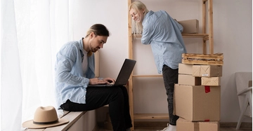 Ein Paar richtet nach dem Umzug die neue Wohnung ein. Während eine Person Regale einräumt, organisiert die andere am Laptop Internet- und Telefonanschluss zwischen Umzugskartons.