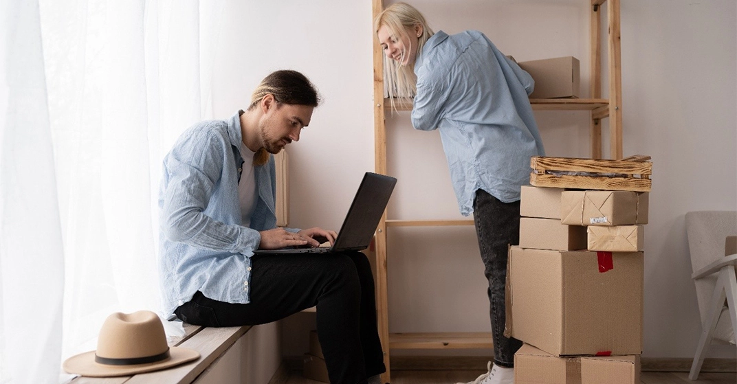 Ein Paar richtet nach dem Umzug die neue Wohnung ein. Während eine Person Regale einräumt, organisiert die andere am Laptop Internet- und Telefonanschluss zwischen Umzugskartons.