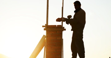 Eine Person mauert einen Ziegelstein auf eine Säule bei Sonnenuntergang. Im Hintergrund ist klarer Himmel zu sehen.
