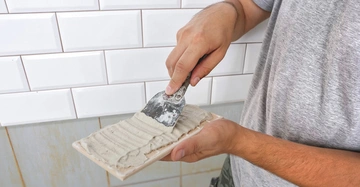 Person streicht Fliesenkleber auf eine Dachziegel mit einem Spachtel vor weißen Fliesen im Hintergrund.