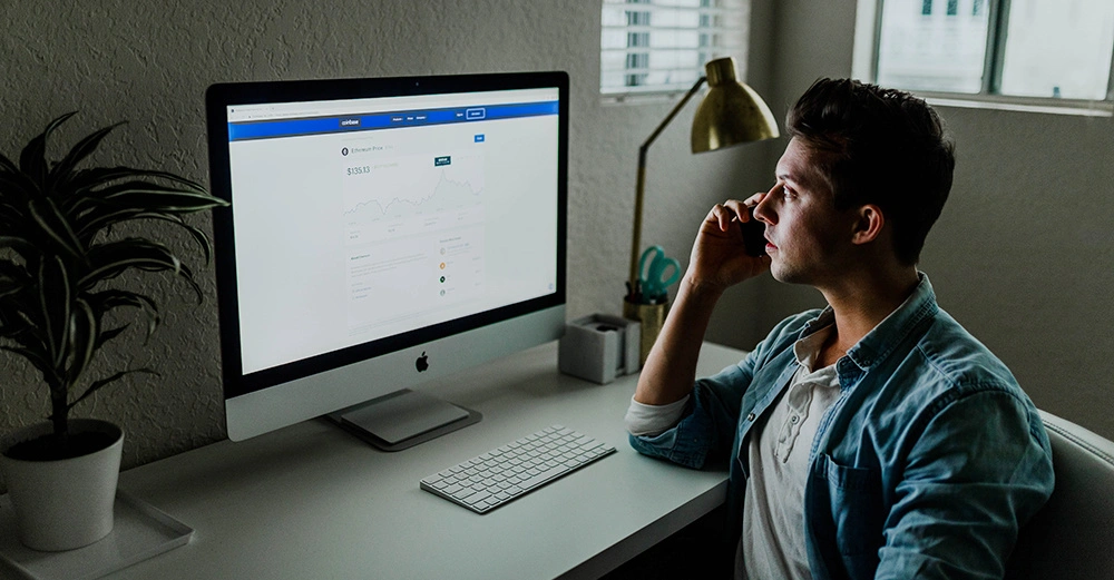 Ein Mann sitzt an einem Schreibtisch und telefoniert neben einem Computerbildschirm mit einer Finanzwebsite in einem gut beleuchteten Raum mit Pflanzen und Schreibtischlampe. Text auf dem Bildschirm: Ethereum Price $135.13.