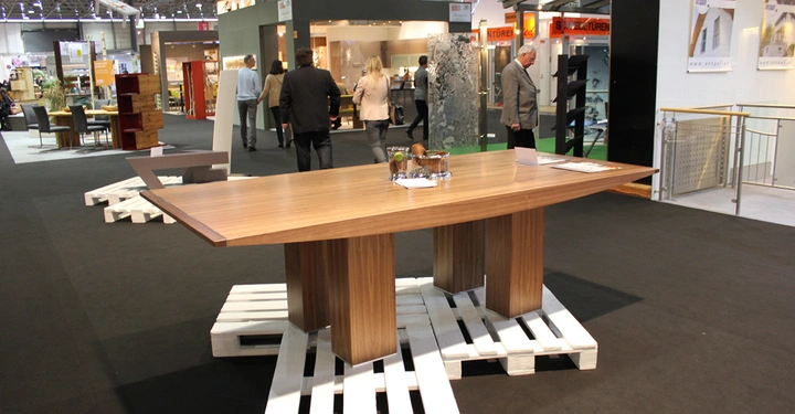 Galerie-Bild 20: Holztisch ruht auf weißen Paletten in einer Ausstellungshalle mehrere Menschen betrachten die ausgestellten Objekte Stände und Möbel umgeben den Tisch.