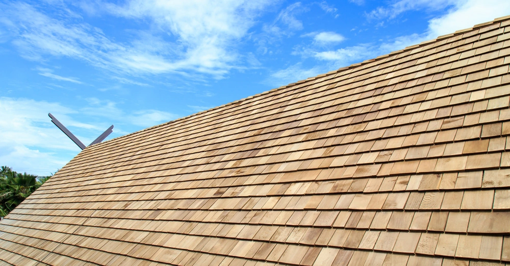 Holzdach mit Schindeln bedeckt in leichtem Winkel unter blauem Himmel mit weißen Wolken zwischendurch und grünen Bäumen am linken Bildrand