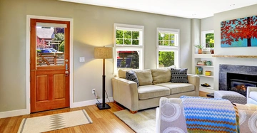 Ein beige Sofa steht im Wohnzimmer vor Fenstern neben einer Holzlampe und einem Kamin. Bunte Dekorationen und Möbel umgeben es bei Tageslicht.