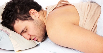 Mann schläft bauchwärts auf einem weißen Kissen und einer blauen Decke in heller Kleidung; naher Bildausschnitt im Schlafzimmer.