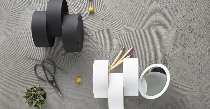 Galerie-Bild 9: Zwei ineinander verschlungene Plastikobjekte halten jeweils Stifte und Blumensträußchen auf einem strukturierten Betonuntergrund, begleitet von einer Schere und einem kleinen Kaktus.