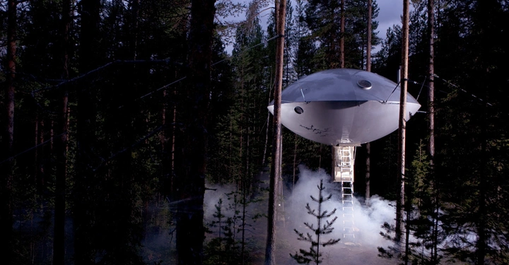 Galerie-Bild 4: Ein UFO-förmiges Baumhaus schwebt zwischen hohen Kiefern in einem nebligen Wald mit einer Leiter, die zum Boden führt.