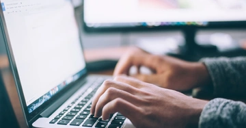 Hände tippen auf einer Laptop-Tastatur in einem Büro mit einem verschwommenen Monitor im Hintergrund.