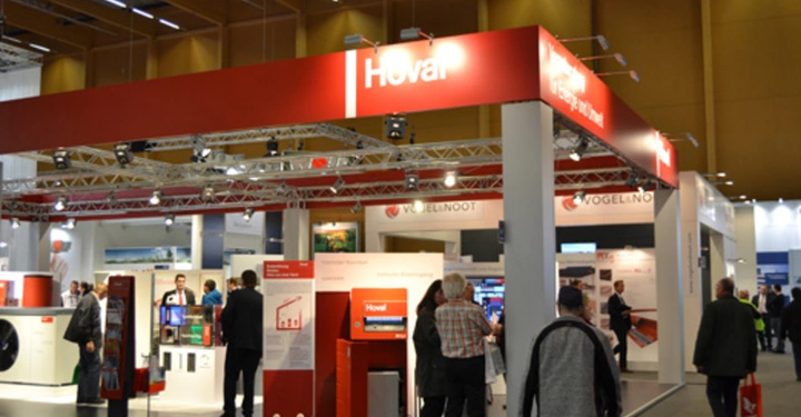 Galerie-Bild 25: Messestand mit dem roten Logo Hoval und VOGEL&NOOT Aussteller und Besucher interagieren Präsentationsflächen beleuchtete Schautafeln und Exponate. Der Innenraum ist modern und professionell gestaltet.