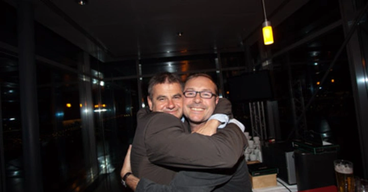 Galerie-Bild 85: Zwei Männer umarmen sich in einem modernen Raum mit Glaswänden im Hintergrund abends zwei Getränkegläser auf einer roten Theke stehend