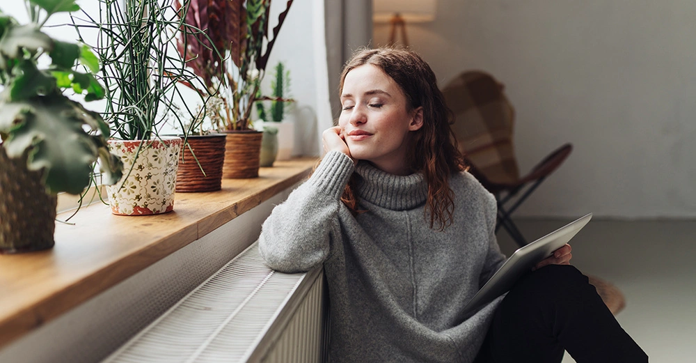 Frau im grauen Pullover hält Tablet sitzt in sonnendurchflutetem Raum auf Fensterbank lehnt Rücken an Heizung mehrere grüne Pflanzen in Töpfen auf Fensterbank Stuhl und Lampe im Hintergrund