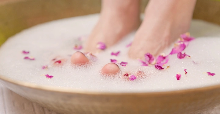 Füße tauchen in eine Schüssel mit schäumendem Wasser, umgeben von Rosenblütenblättern auf einer hellen Holzoberfläche.
