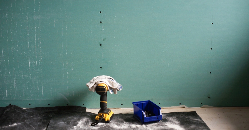 Akkuschrauber mit Tuch bedeckt steht auf einem Boden mit schwarzer Abdeckung neben einem blauen Behälter vor einer grünen Wand