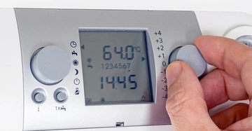 Hand dreht den Knopf an einem modernen Thermostatgerät Temperaturanzeige 64.0°C Uhrzeit 14:45 Symbole und digitale Anzeige auf grauem Hintergrund.