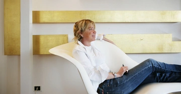 Frau sitzt entspannt auf einem modernen weißen Stuhl hält einen Stift lächelt und blickt zur Seite im Hintergrund weiße Wand mit horizontalen goldenen Streifen.