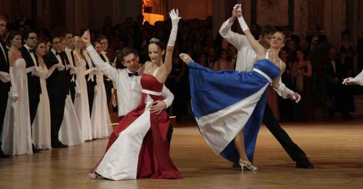 Tanzpaare in formeller Kleidung führen einen Walzer auf einem Parkettboden auf, während Zuschauer in einem eleganten Ballsaal zuschauen.