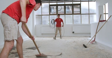 Zwei junge Männer, beide in einem roten T-Shirt und einer kurzen beigen Arbeitshose, bereiten den Estrich-Untergrund vor. Einer arbeitet mit einer Stoßscharre, der andere wickelt ein langes Kabel ab. Schaufel und Besen lehnen an der Wand.