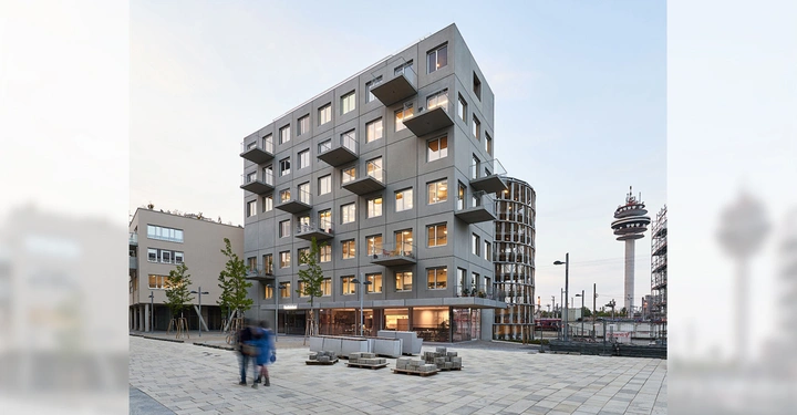 Sechsstöckiges Gebäude mit Balkonen steht auf einem gepflasterten Platz mit Bäumen. Zwei Personen gehen im Vordergrund. Ein Fernsehturm befindet sich im Hintergrund zusammen mit einem zweiten Gebäude.