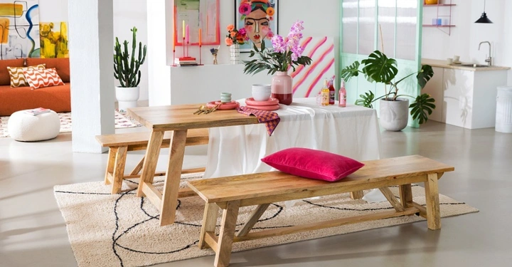 Farbenfroher Essbereich mit Holztisch, Bänken und pinken Akzenten. Im Hintergrund bunte Wandkunst, ein Kaktus und eine Monstera. Ein moderner, eklektischer Raum mit hellem Boden und viel Tageslicht.