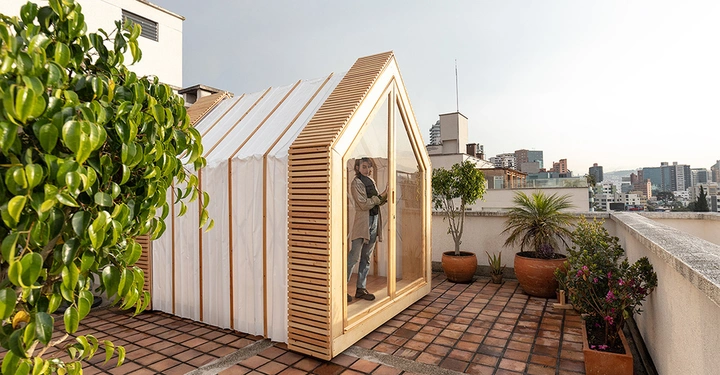 Galerie-Bild 3: Junge Frau steht hinter Glasschiebetür vom Mini-Falthaus IWI, auf städtischer Dachterrasse mit großen Kübelpflanzen, Sonnenuntergang