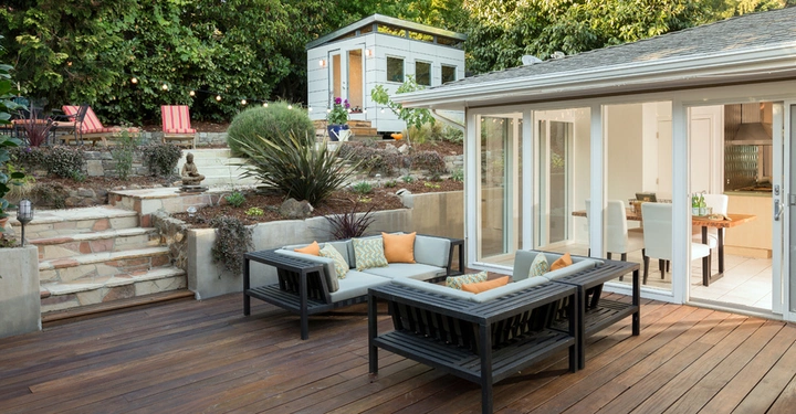 Gartenmöbelgruppe steht auf einer Holzterrasse vor einem Haus und Sitzkissen sind vorhanden. Es gibt eine Treppe, die zu einem höher gelegenen Bereich mit einem Gartenhaus und weiteren Sitzbereichen führt.