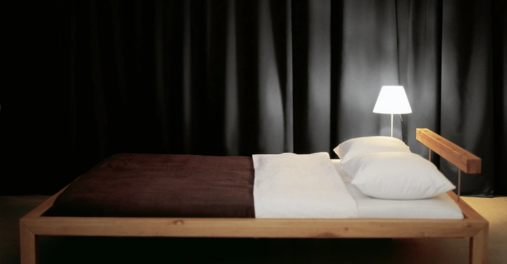 Galerie-Bild 6: Holzbett mit brauner und weißer Bettwäsche steht in modernem Schlafzimmer mit schwarzen Vorhängen und einer eingeschalteten Tischlampe auf dem Nachttisch.