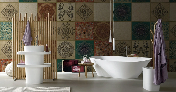 Weiße Badewanne und Waschbecken stehen still in einem Badezimmer mit bunten Wandfliesen in verschiedenen Mustern und Farben. Handtücher hängen an Ständern; Teppich und Hocker ergänzen die Einrichtung.