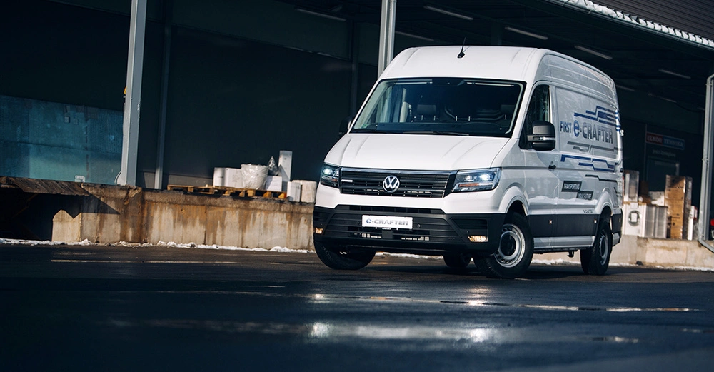 Weißer Volkswagen Transporter steht auf nasser Fahrbahn vor einer Lagerhalle im Industriegebiet. Auf dem Fahrzeug steht der Text: e-CRAFTER FIRST INNOVATIVE mehr bewegen.