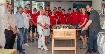 20 Menschen versammeln sich rund um einen Tischtennistisch, viele in Hartl-Rot gekleidete Lehrlinge, dazu 3 Ärztinnen bzw. Pflegerinnen und Vertreter von Hartl Haus lächeln in die Kamera. Einige mit Händen am Wuzzler