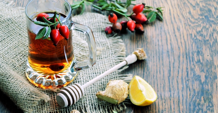 Glaskrug mit Tee steht auf Sackleinen Tuch neben frischen Hagebutten Honiglöffel Ingwerstücken und Zitronenscheiben auf rustikalem Holztisch.