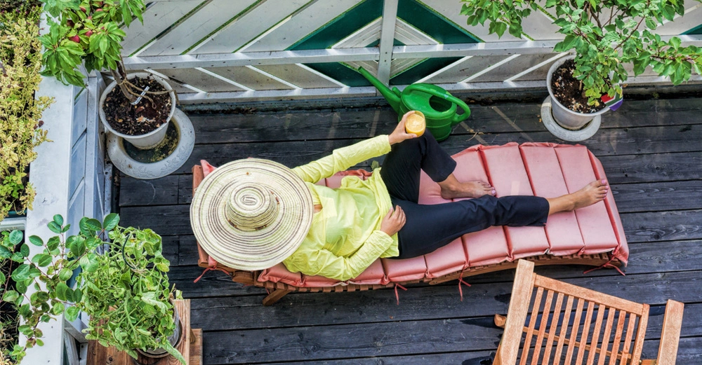 Frau mit Sonnenhut entspannt am Balkon auf einem Liegestuhl mit rot gepolsterter Auflage. Sie hält ein Glas Orangensaft in den Händen, um sie herum Topfpflanzen.