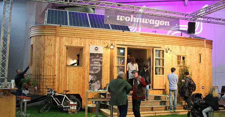Galerie-Bild 5: Holzwagen mit Solarzellen auf dem Dach Menschen gehen hinein oder stehen davor grüne Wiese Umgebung Werbebanner mit dem Text wohnwagon ein Plakat sagt AUTARK und NATÜRLICH leben Tisch auf der linken Seite mit Pflanzen und Fahrrad