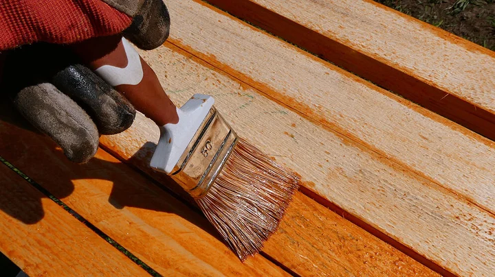 behandschuhte Hand streicht mit kleinem, breitem Pinsel eine Holzlasur auf dünne Latten von vielleicht einem Gartentisch