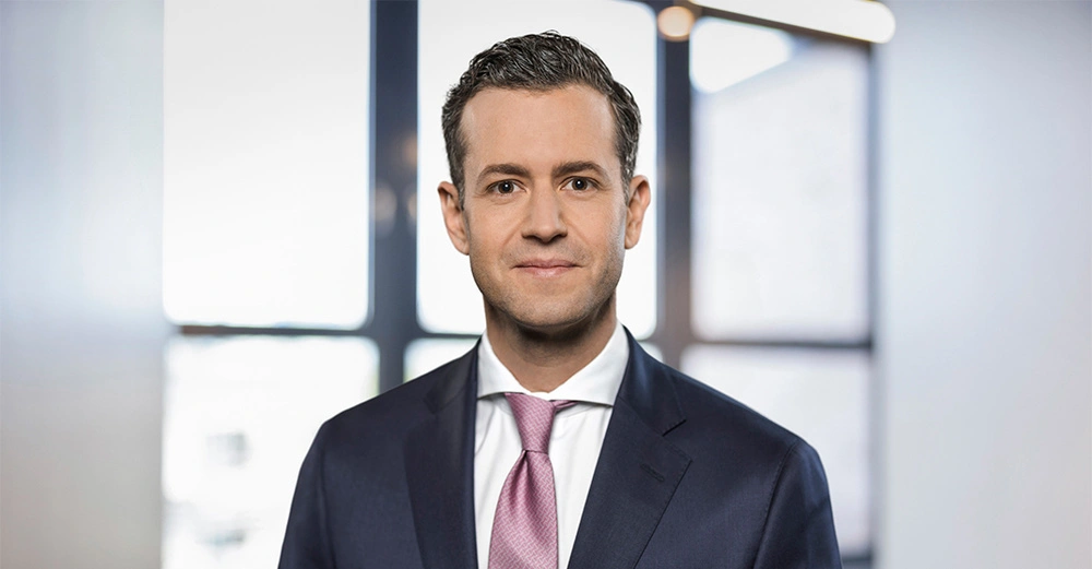 Ein Mann trägt einen Anzug mit Krawatte und lächelt. Im Hintergrund sind verschwommene Fenster, die ein modernes Büro andeuten.