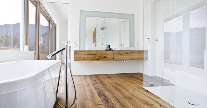 Badewanne steht neben einem freistehenden Wasserhahn in einem modernen Badezimmer mit Holzboden. Ein großes Fenster bietet Blick auf die Berge. Ein Holzwaschtisch mit Spiegel hängt an der Wand.