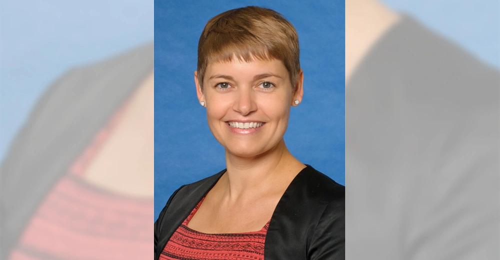 Frau mit kurzem Haar lächelt in die Kamera trägt ein schwarz-rotes Oberteil und eine schwarze Jacke steht vor einem blauen Hintergrund