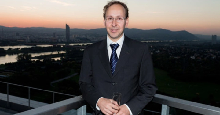 Galerie-Bild 46: Ein Mann im Anzug hält ein Glas in den Händen und steht auf einem Balkon. Im Hintergrund ist eine städtische Landschaft mit Fluss und Hügeln bei Sonnenuntergang zu sehen.